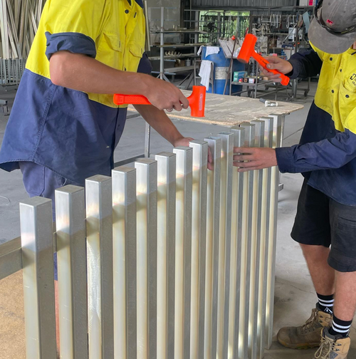 Two workers in high-visibility jackets installing aluminum end caps into the ends of metal frames in a workshop.